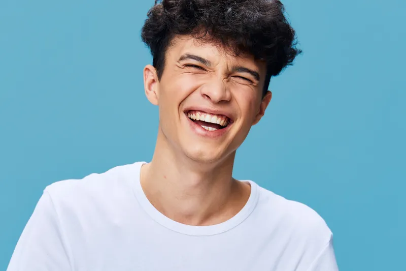 Laughing man with curly hair wearing white t-shirt against blue background, showing joyful expression and natural happiness in simple casual style.