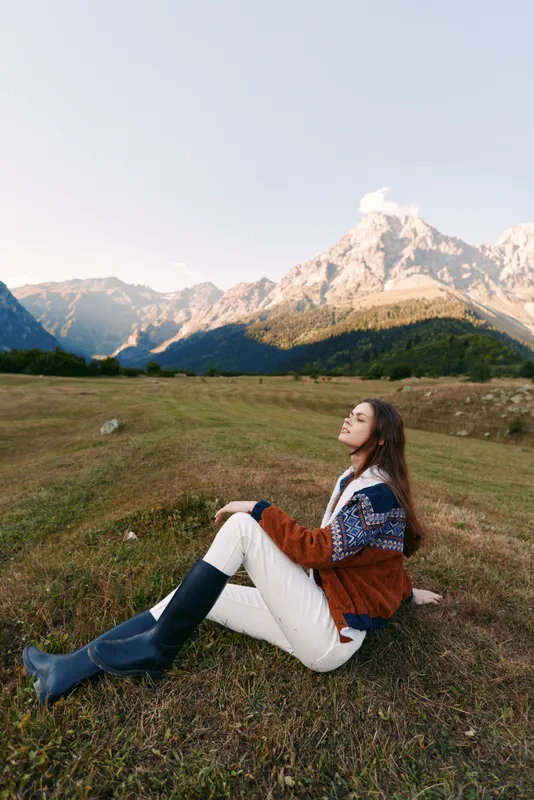 Woman in sweater and boots sitting in meadow, mountains landscape behind, nature scene of autumn travel and relaxation. Young portrait enjoying peaceful, serene outdoor moment.