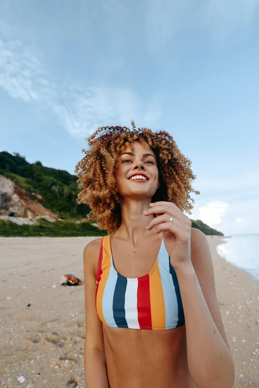 Young woman, beach, curly hair, smiling in summer, wearing striped swimsuit, on sandy shore by ocean, sunny outdoor day, hand near face, relaxed happy mood, breezy wind, gentle waves nearby