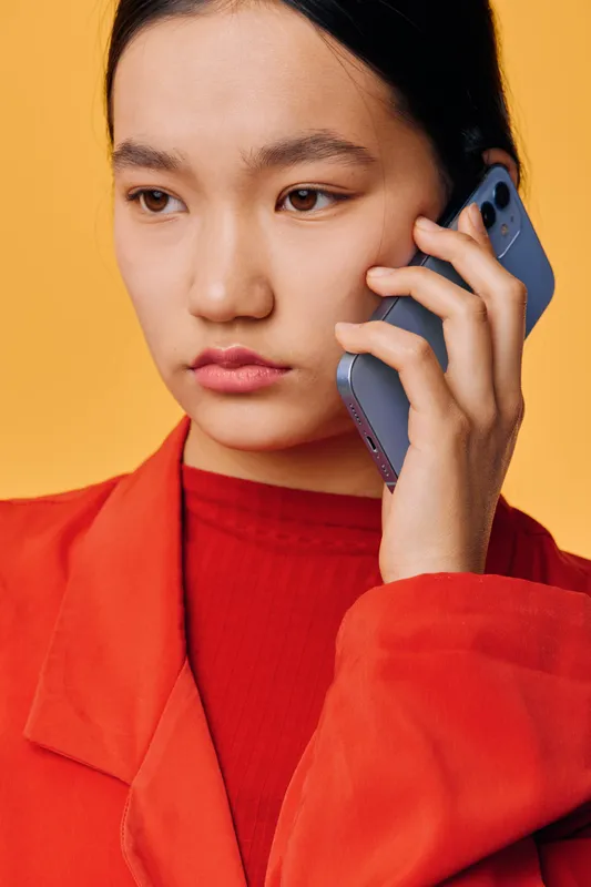 Young woman in red jacket speaking on smartphone with serious expression, isolated on warm yellow background. Closeup portrait, communication, technology, and connection concept.