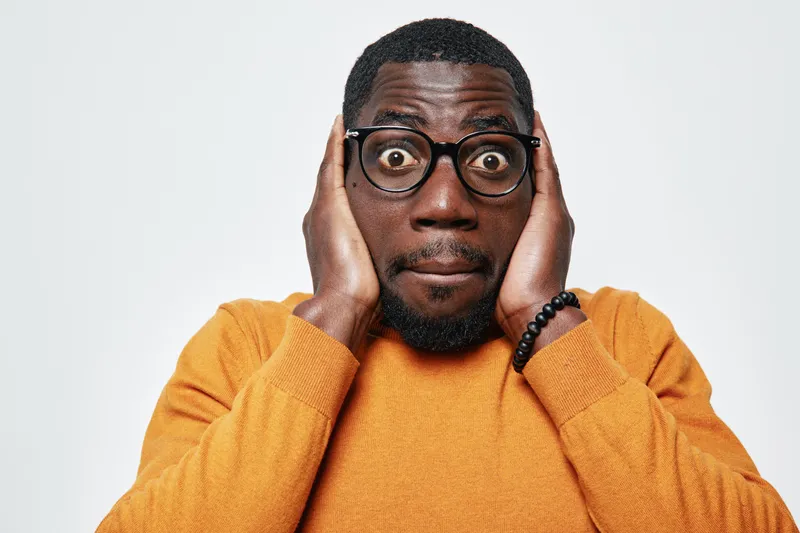 African man with dark skin and glasses wearing orange sweater covering ears with hands, expressing surprise or shock on white background. Casual portrait with emotion and reaction.