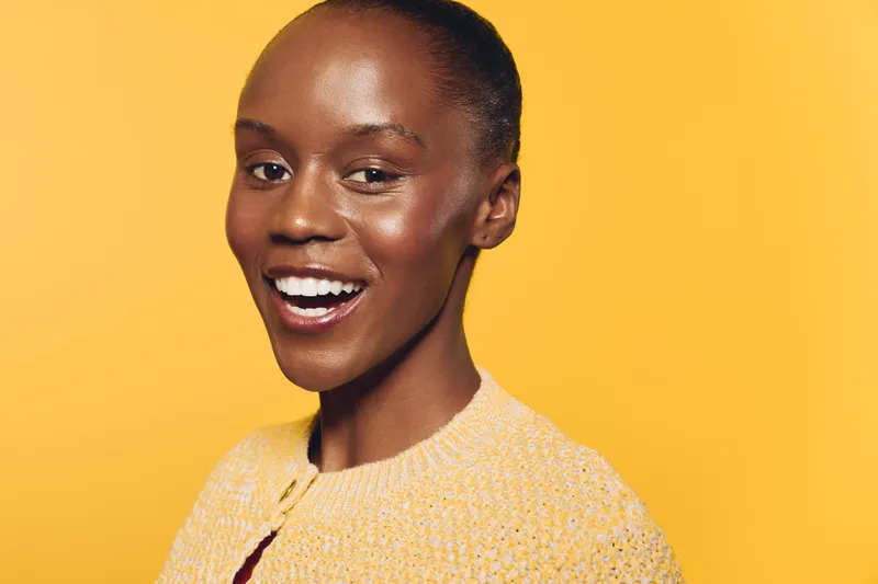 Happy smiling African woman with dark skin wearing yellow sweater on orange background. Close-up portrait with natural makeup, showing positive emotion and joyful mood in studio lighting.