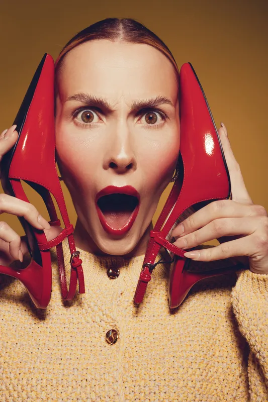 Surprised woman with red lipstick holds red high heel shoes near face, wearing yellow sweater. Studio portrait with expressive emotion and fashion concept on beige background.