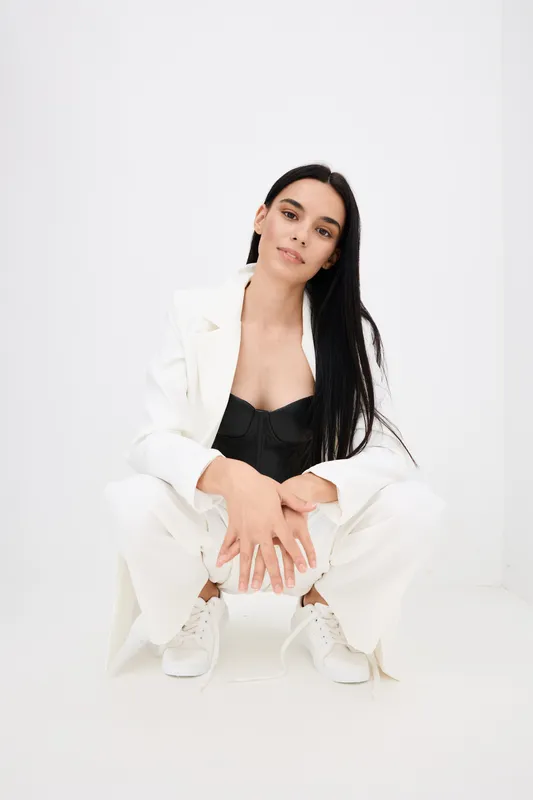 young woman posing in white wide-leg trousers and blazer with black top, long dark hair, casual modern look, minimalistic white background, confident expression