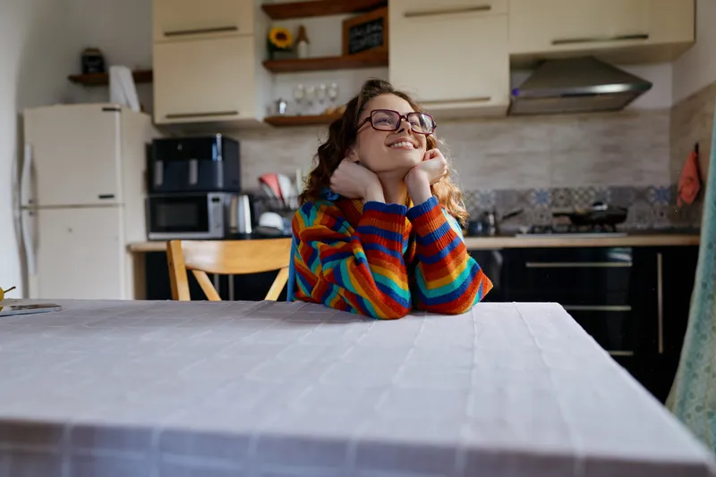 Casual woman at home in a cozy kitchen, colorful striped sweater, dreamy smile, relaxed pose, warm natural light, domestic setting, everyday moment, content mood, reflective pause, quiet serenity