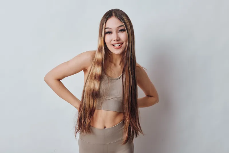 Young woman with long hair wearing beige sportswear smiling and posing with hands on hips in front of white background. Fitness, casual style and confident expression.