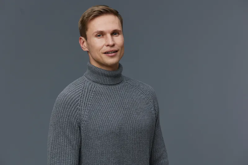Full body portrait of a cheerful young Caucasian man with short light brown hair, wearing a cozy gray turtleneck sweater. He smiles gently while looking at the camera against a plain gray background. 