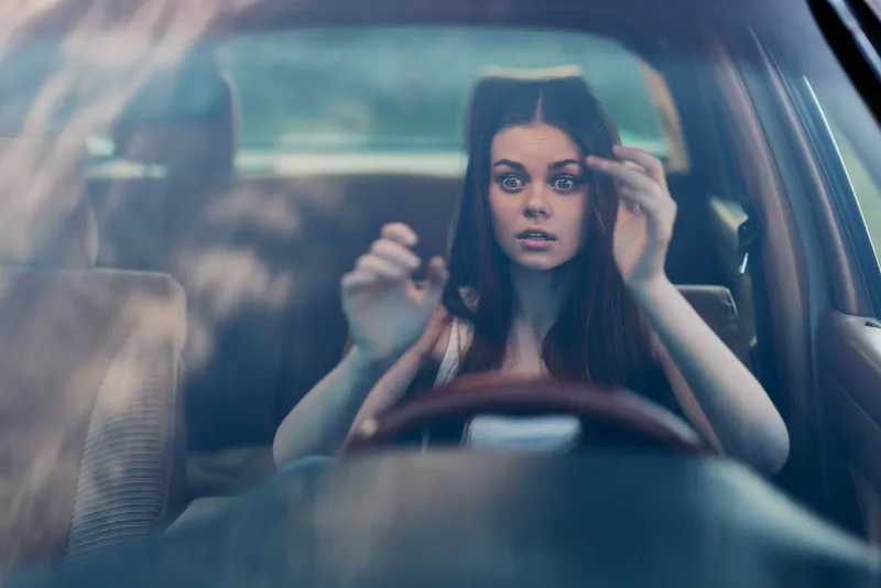 Young woman with long brown hair looking surprised while driving a car, showcasing a vulnerable emotional moment in a natural setting.