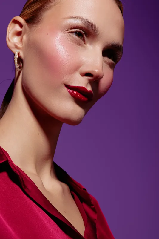Close-up portrait of a young woman with glowing skin and red lipstick wearing a red blouse against a purple background. Concept of beauty, makeup, fashion, and skincare.