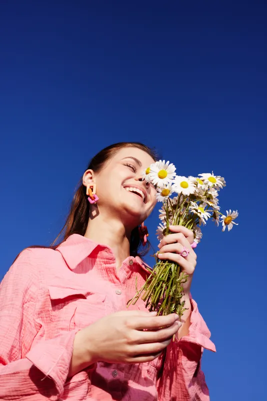 Young woman in pink outfit holding daisies against a blue sky, expressing joy and warmth, perfect for spring or summer themes