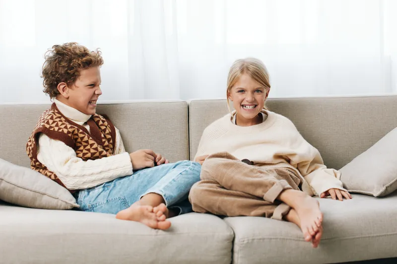 Two children sit on a sofa, mid playful quarrel with tense expressions and grins, wearing cozy sweaters in a bright, warm living room. A candid moment of childhood rivalry.