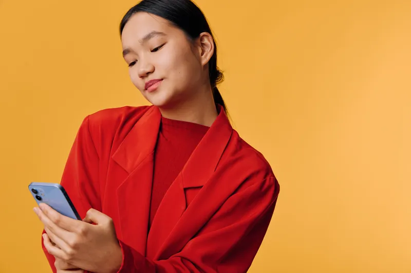 Young woman in red jacket holding and looking at smartphone with slight smile on yellow background. Female using mobile phone for communication or social media with casual style.