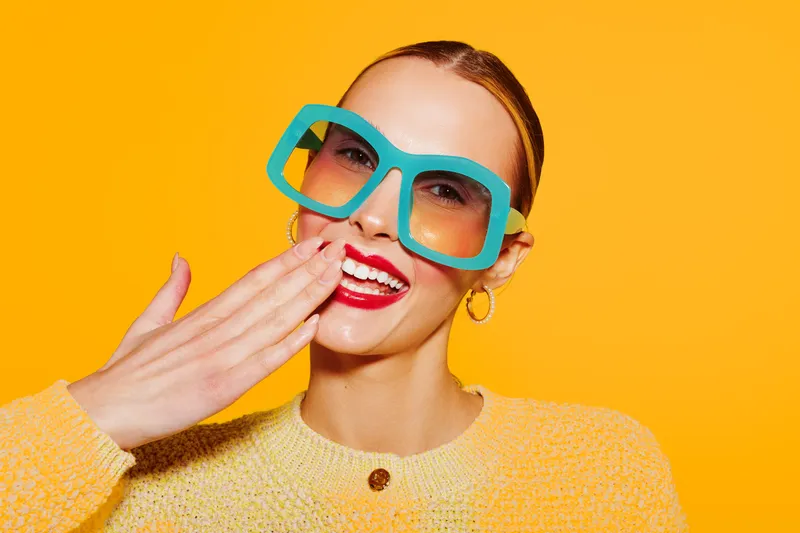 Smiling woman with large blue sunglasses and red lipstick covers mouth with hand on bright yellow background. Concept of fashion, summer style, and joyful mood.