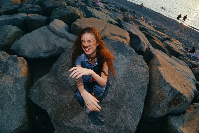 happy woman embracing modern boho-western denim street style sitting on large rocks at sunset with warm film color tones, lifestyle outdoor coastal scene