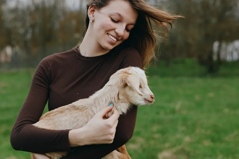 Young woman hugs a friendly goat kid in a green field, enjoying a casual outdoor moment with a warm smile, natural light, and rural lifestyle vibe, suitable for family, pets, and countryside stories