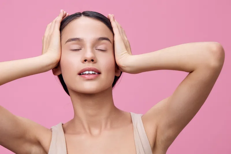 Young woman with closed eyes expressing tranquility while holding her head, against a soft pink background, embodying relaxation and calmness in a minimalist style