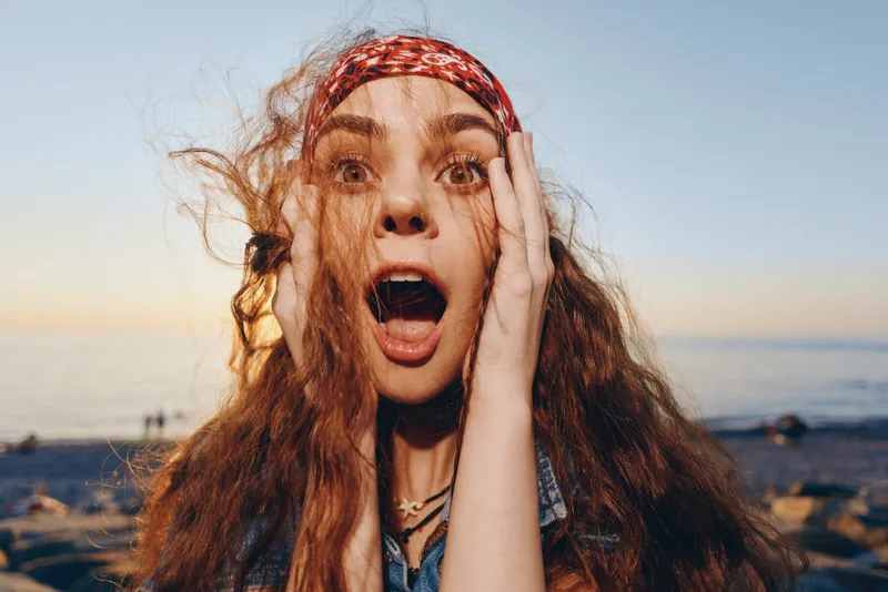 surprised woman with curly hair in modern boho-western denim street style expressing shock outdoors at sunset, film color vibes, lifestyle emotion