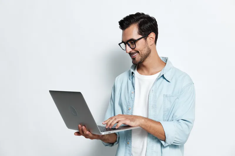 Smiling young man wearing glasses and denim shirt, using laptop computer while standing against plain light background. Stylish modern lifestyle concept.
