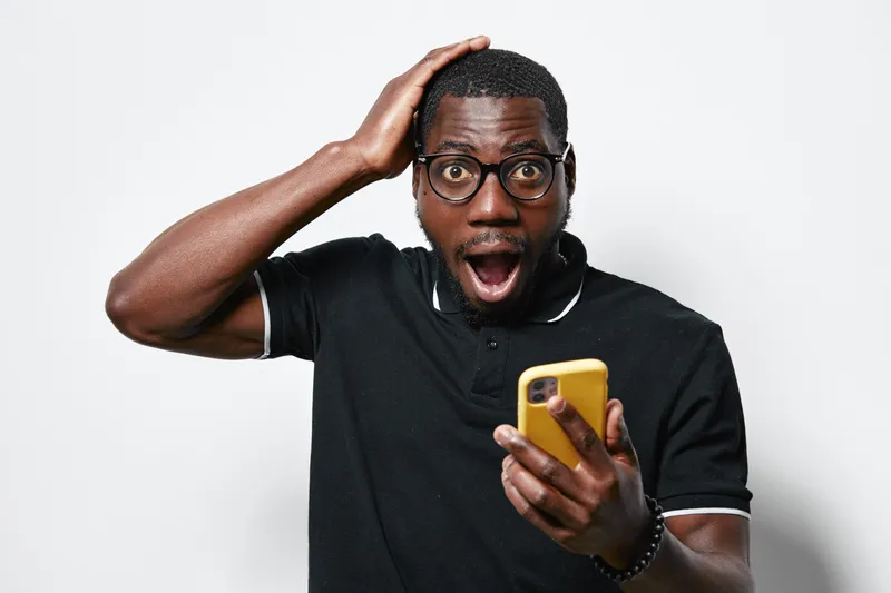 African American man with dark skin and glasses holding yellow smartphone with surprised expression on white background. Casual black polo shirt and short hair.