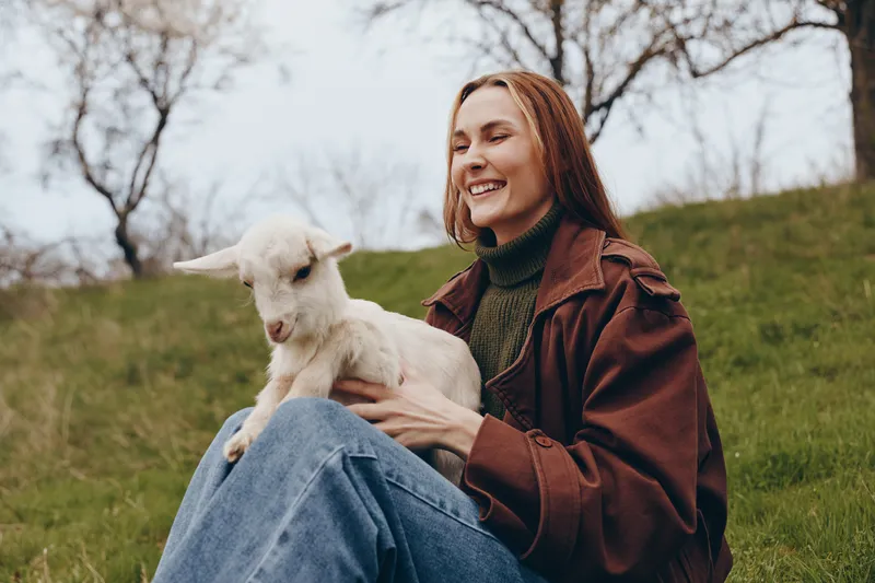 Smiling woman in a casual jacket holds a goat kid outdoors on a grassy hill, capturing a warm lifestyle moment with animals, nature, and gentle companionship.