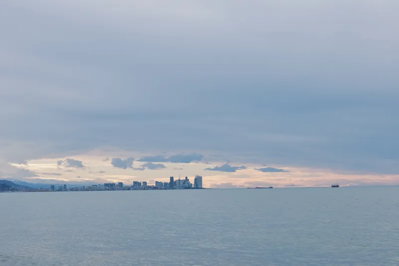 City skyline across calm water under overcast sky, distant buildings on the horizon, tranquil seascape with soft light and distant ship
