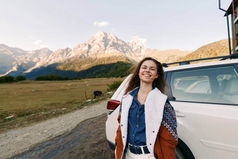Woman, car, mountains, travel, smile, outdoors, road, adventure: Happy young woman standing by a white SUV on a rural mountain road, smiling during a scenic outdoor road trip with towering peaks.