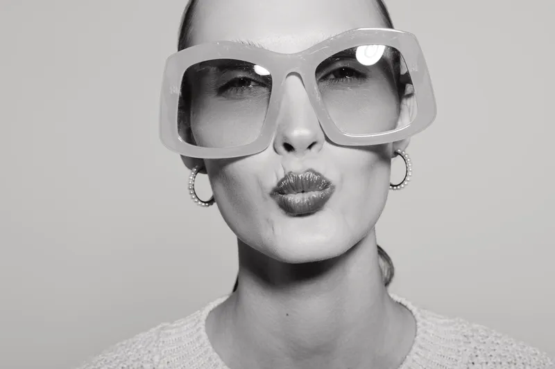 Portrait of a Caucasian woman with fair skin wearing large transparent glasses and earrings, making a kiss face. Studio lighting emphasizes facial features and fashionable accessories.