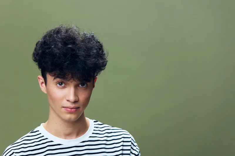 young man with curly hair wearing striped shirt looks confidently against olive green background in studio portrait, casual style, natural light