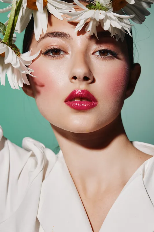 Close-up portrait of a woman with vibrant makeup and glossy pink lips wearing white clothes and daisies around her head. Fashion beauty concept with creative floral styling and soft lighting.