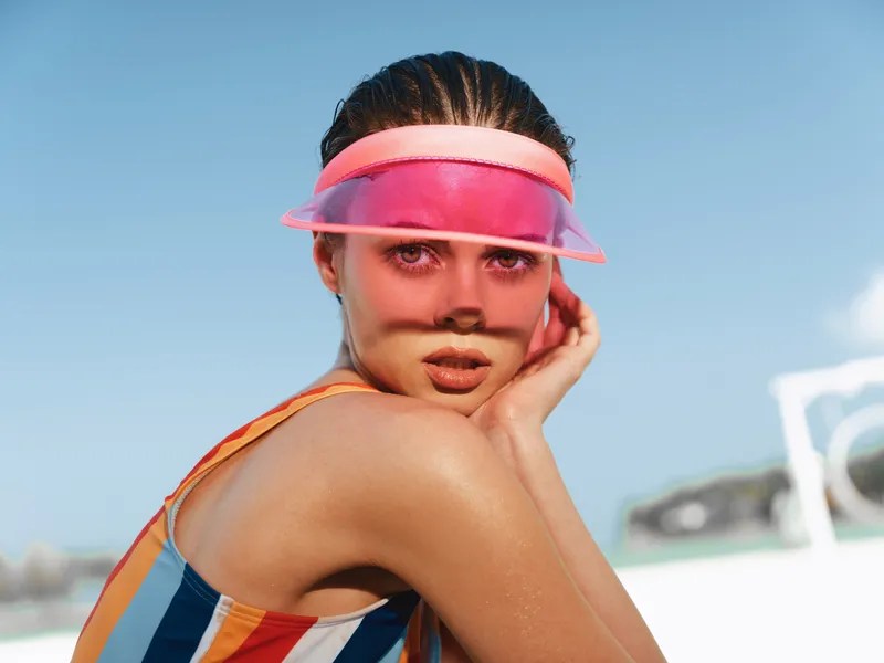 Portrait of a sunlit model wearing a bright pink visor and striped swimsuit, looking toward the camera with a soft, calm expression against a clear blue sky.