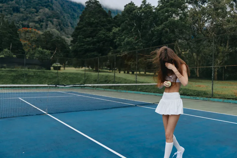 Young female tennis player in a white skirt, standing on a blue court, holding a tennis ball with a playful expression, surrounded by green trees and mountains