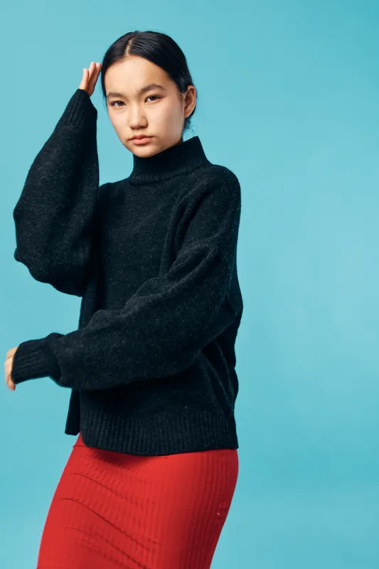 Young woman posing in black sweater and red skirt, standing against blue background in studio. Fashion model with serious expression, casual style, and long dark hair for portrait photography.