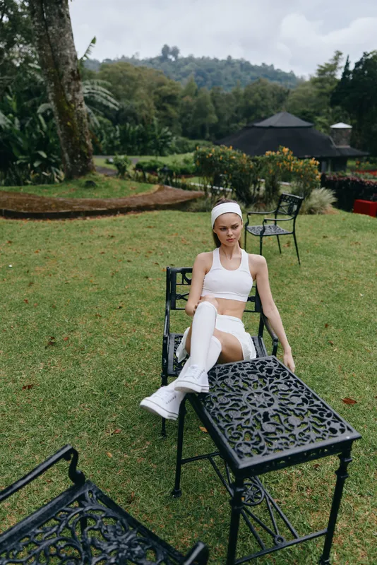 A fashionable young woman in sporty white activewear relaxes outdoors in a lush green garden, embodying wellness and fitness lifestyle