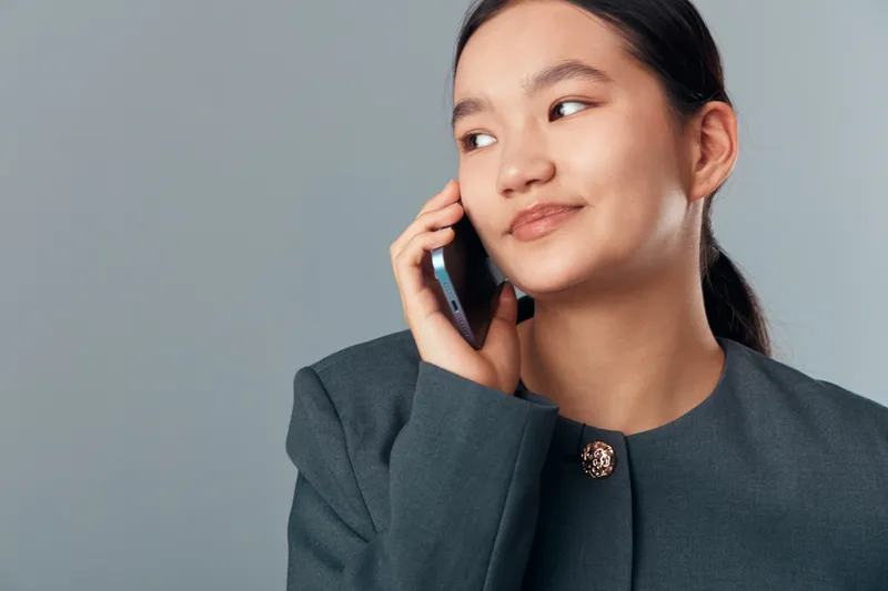 Young woman speaking on smartphone with thoughtful expression wearing formal jacket, communication and technology concept, business or casual style with gray background.
