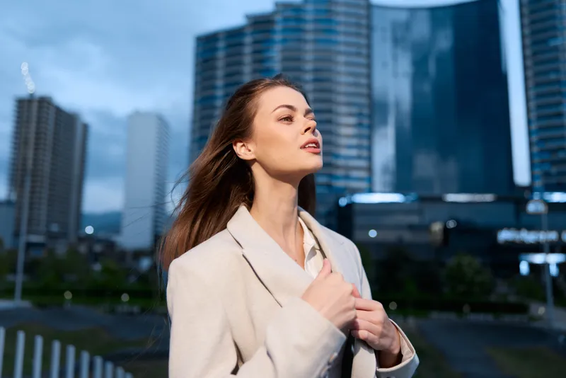 Confident businesswoman gazing upwards in a modern urban setting, embodying empowerment and ambition, dressed in a stylish coat against a dynamic city backdrop.