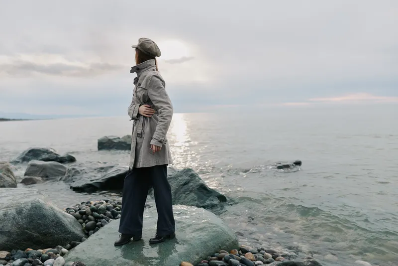 A person in a coat and hat stands on rocky shore looking at calm sea under cloudy sky at sunset. Concept of solitude, nature, and peaceful outdoor moment by water.