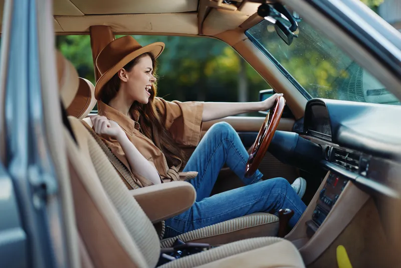 Cheerful young woman with a stylish hat enjoying a road trip, with casual attire and an energetic expression in a vintage car interior.