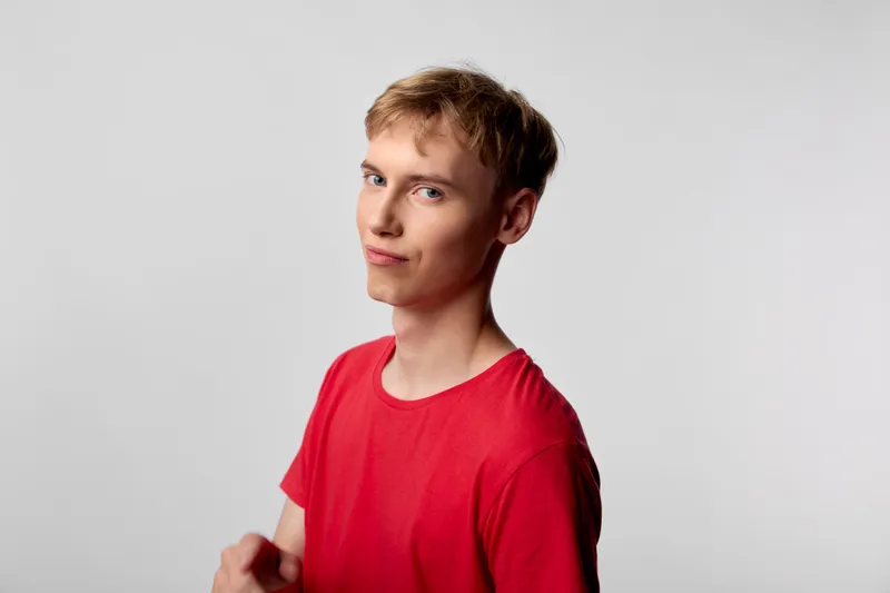 Young man in a red shirt with a subtle smirk stands in a studio against a neutral backdrop, delivering a casual lifestyle image suitable for fashion, menswear, or social media campaigns.