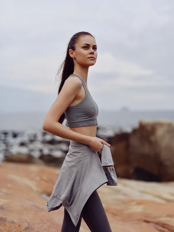 Fit young woman in gray activewear jogging along the rocky beach, embodying health and vitality with a determined expression, ideal for fitness lifestyle imagery