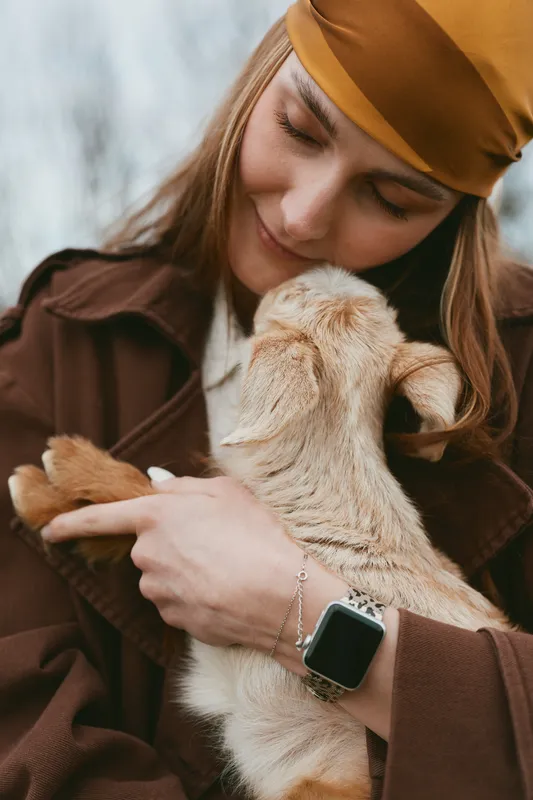 Young woman in brown coat gently holds a baby goat close to her chest outdoors, showing affection and warmth in a candid lifestyle moment.