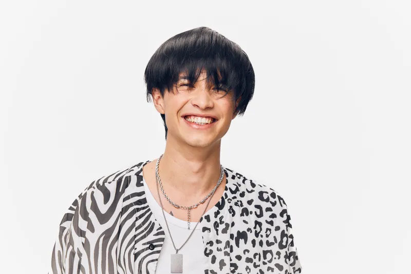 young man smiling happily wearing patterned shirt and layered silver necklaces, isolated on white background with trendy hairstyle and expressive eyes