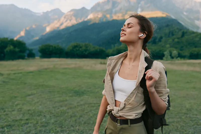 Woman enjoying outdoor lifestyle in a sunlit meadow, mountains in the background, casual backpack fashion, embracing nature and relaxation.