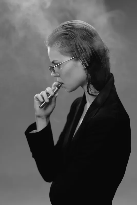 Black and white portrait of a woman vaping with smoke, wearing glasses and a dark jacket, modern and stylish.