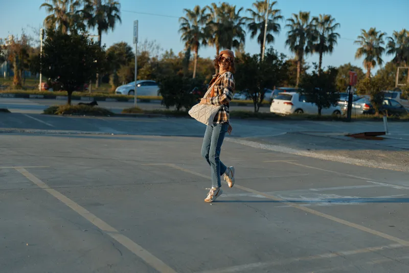 Woman jump in golden light, tote and sunglasses, jeans in a parking lot by palm trees. Candid lifestyle shot with authenticity, emotional storytelling, mindful living and glow.