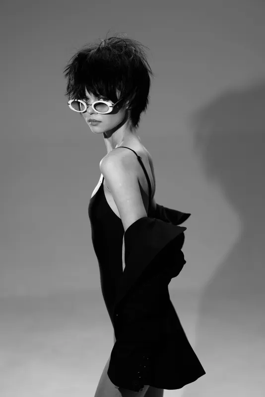 Black and white portrait of a stylish woman with short hair wearing sunglasses and a loose jacket, showcasing modern fashion and confident attitude.