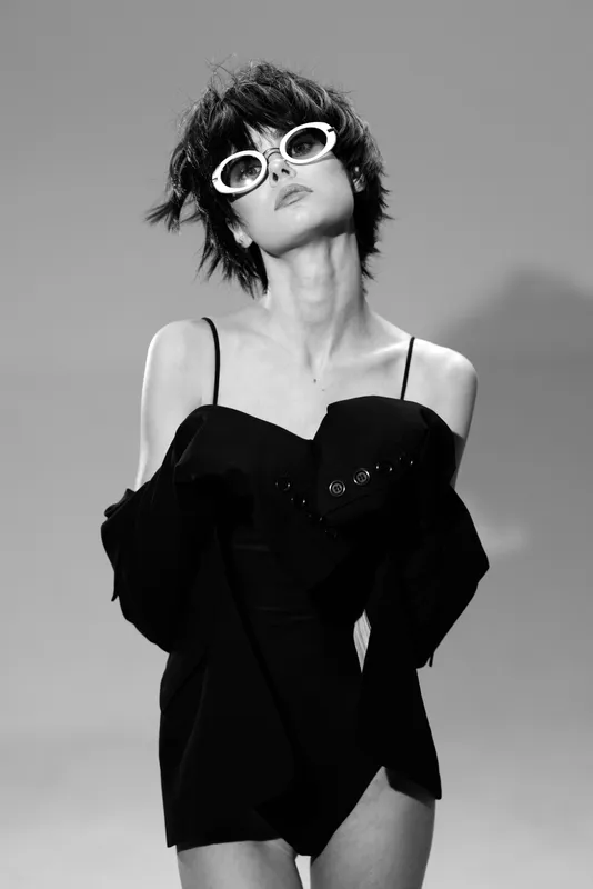 Stylish woman in black dress wearing white sunglasses, posing confidently with short hair in a black and white studio setting.