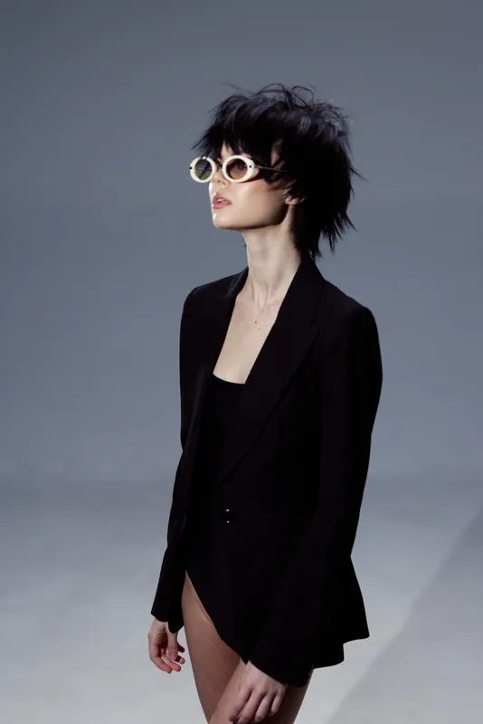 Stylish woman with short dark hair wearing round sunglasses and black jacket posing against gray background.