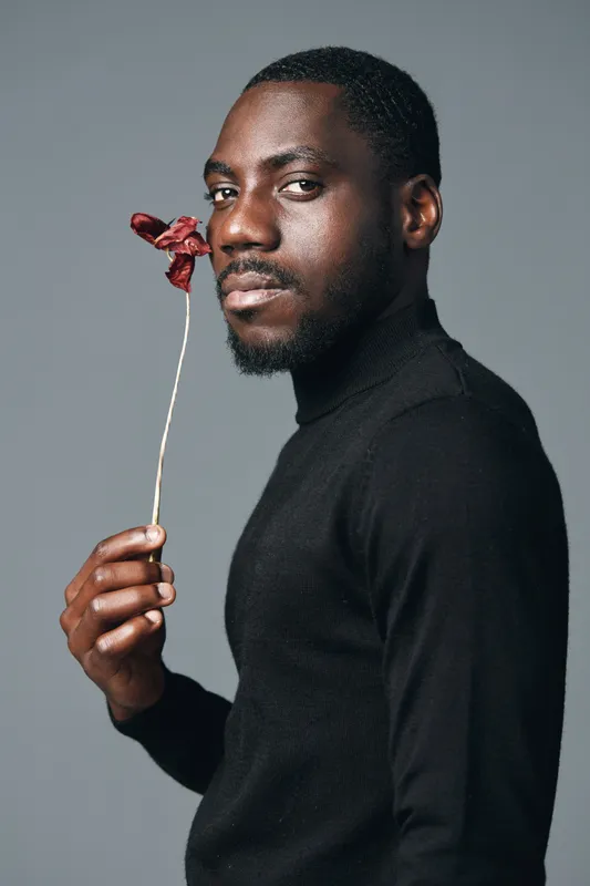 Black man with dark skin holding dried red flower near face in creative visual designer style. Portrait with serious expression and black turtleneck on gray background.