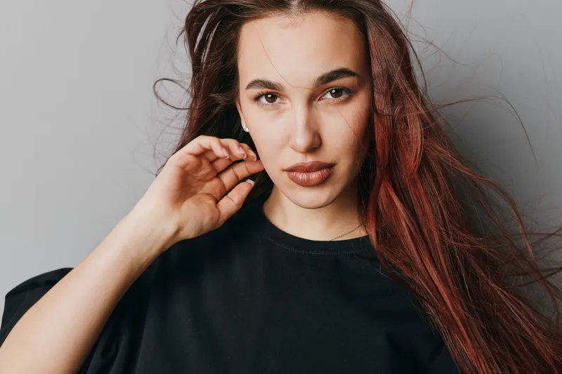 Young woman with long red hair wearing black shirt posing with hand near face against plain background. Portrait, beauty, confident expression and casual style for fashion or lifestyle.