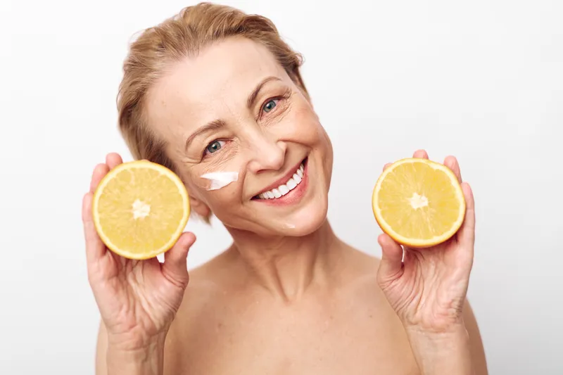 Smiling mature woman with short hair holds two orange halves near face with cream on cheek, isolated on white background. Concept of natural skincare, healthy lifestyle, and beauty care.